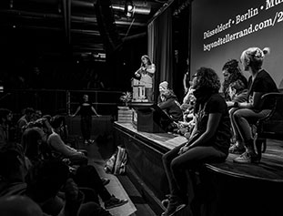 photo en noir et blanc prise sur le coté de la scène, permettant de voir le public sur le coté gauche et la scène sur le coté droit avec des conférenciers dessus