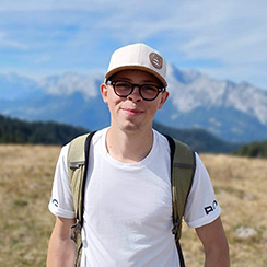 Avatar de Justin, photo prise en plein air; homme binoclard portant un tshirt blanc et une casquette blanche, il porte également sur son dos un sac à dos de randonnée; il a l'air jovial et se situe sur la plaine d'une région montagneuse